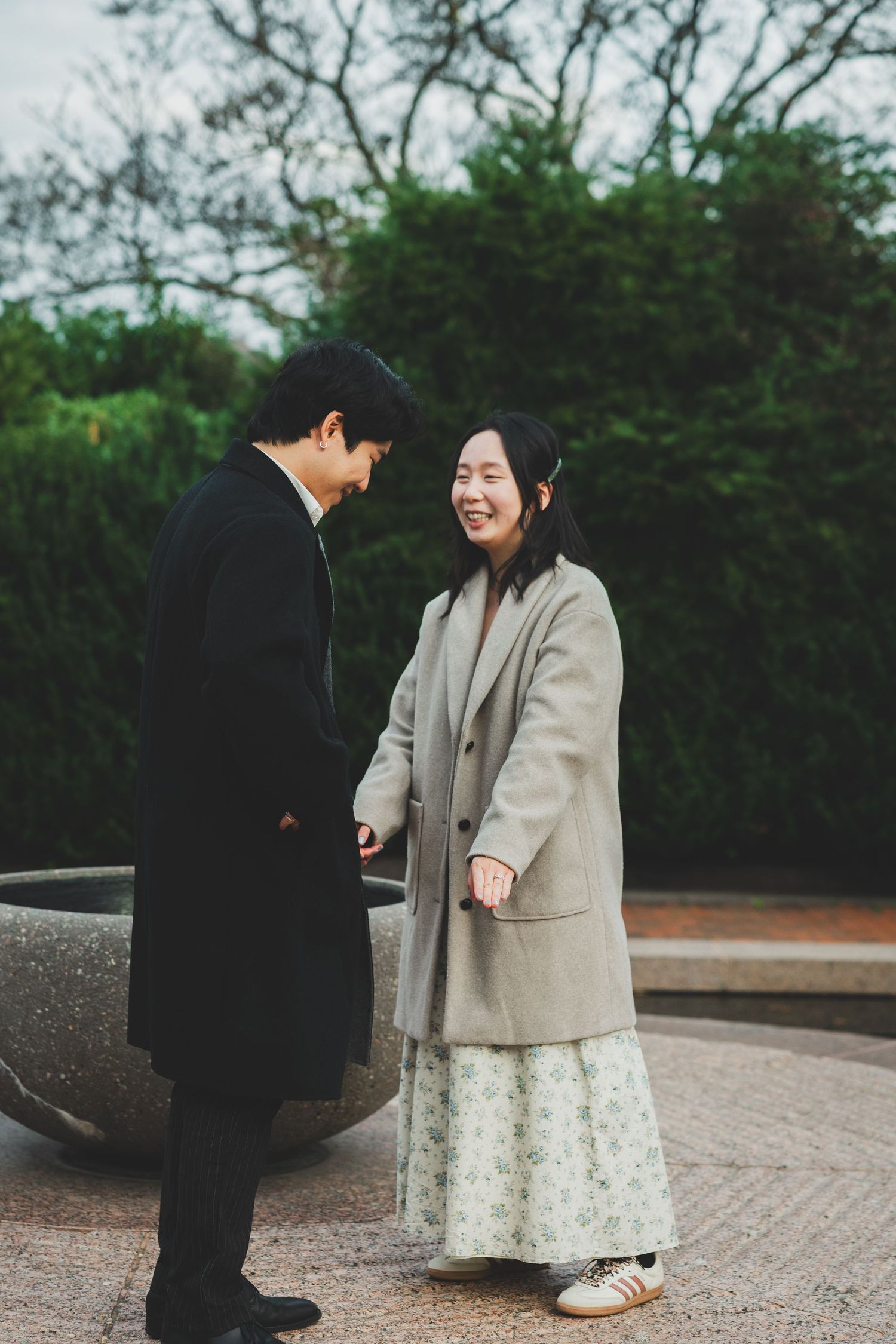 A smiling couple in winter coats stands together in a park, sharing a warm moment near a stone fountain.