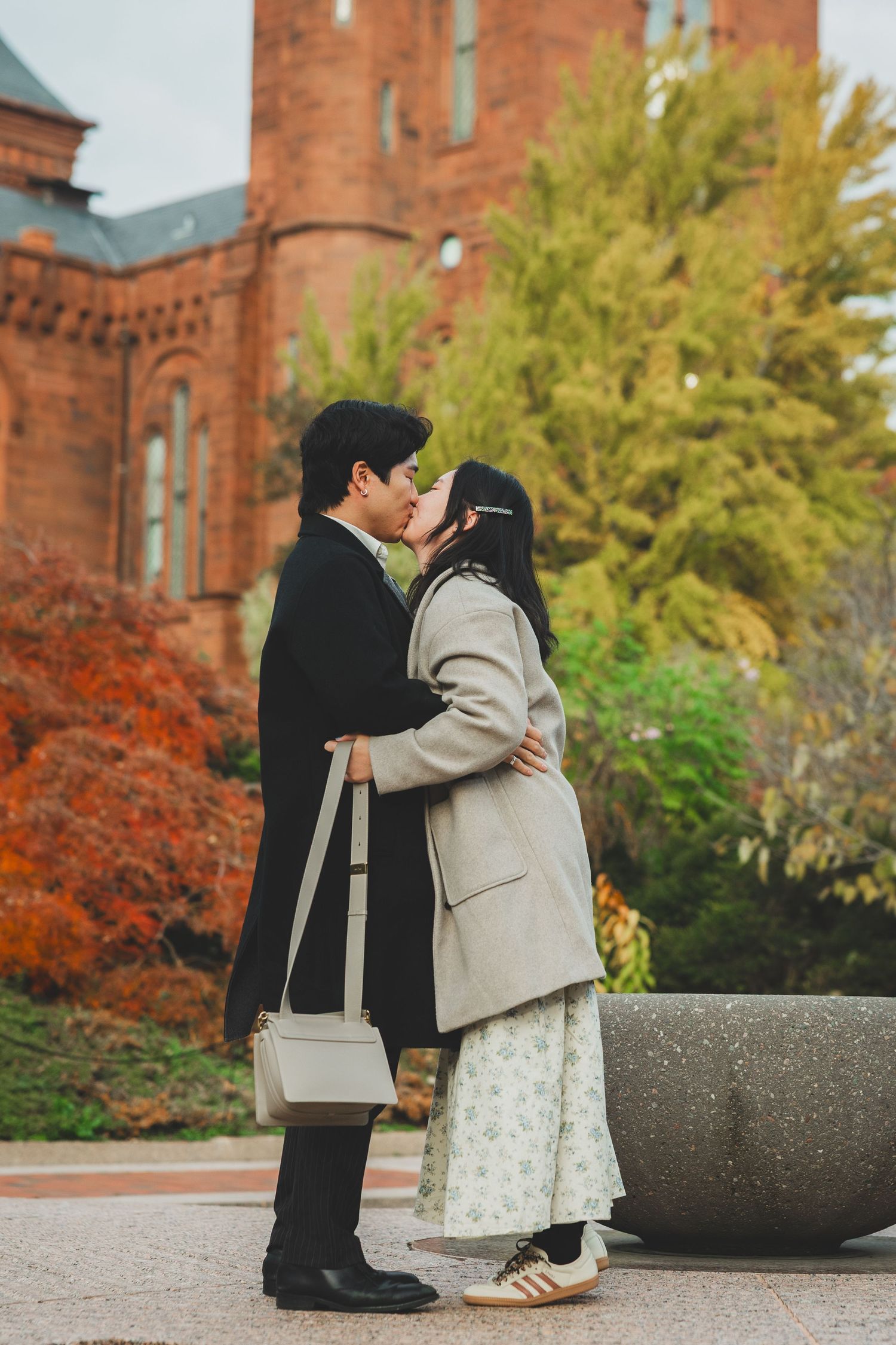 A couple shares a romantic kiss outdoors in autumn, dressed in elegant coats near a red brick building.