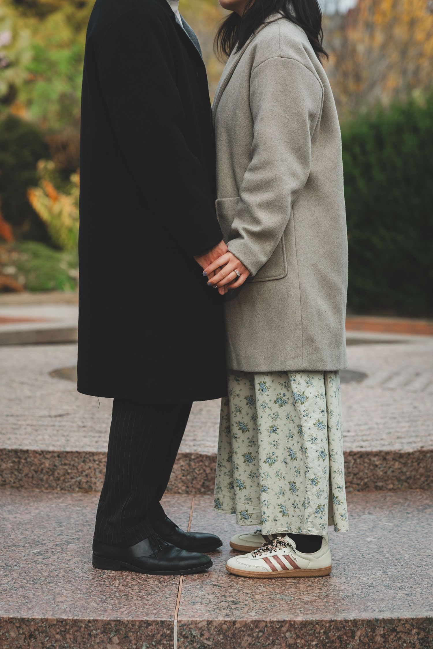 Couple holding hands in autumn park, wearing long coats, floral skirt, and Adidas sneakers on marble steps.