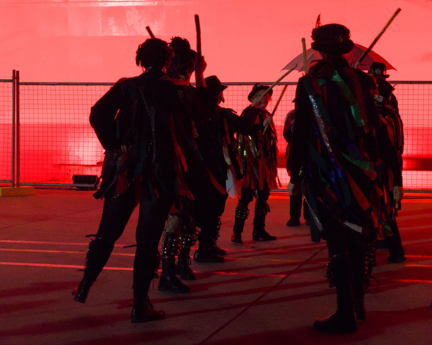 Performers in dark costumes with colorful ribbons stand in red-lit venue during dramatic cultural dance performance.