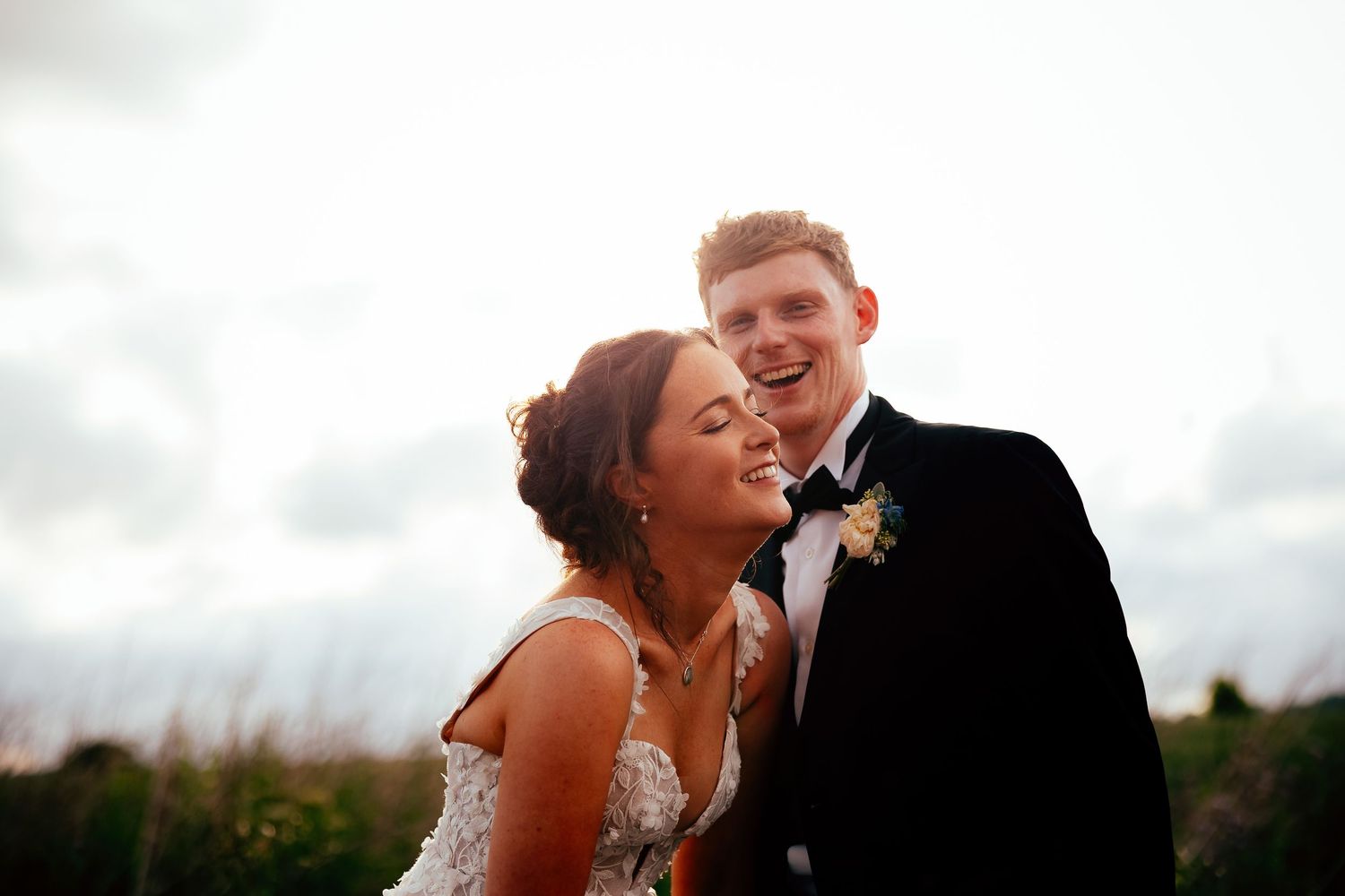 Bride and groom share a romantic moment kissing outdoors with golden backlight and cloudy sky at their wedding.