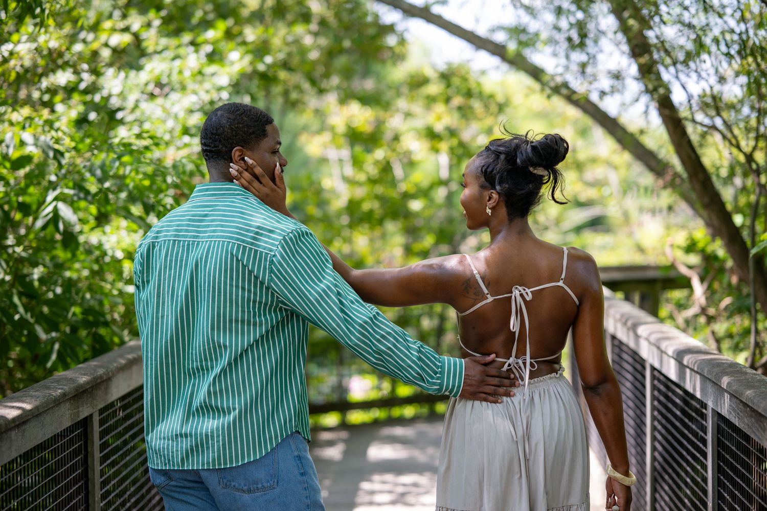 A couple walks hand-in-hand on a wooden bridge surrounded by lush green trees on a sunny day.