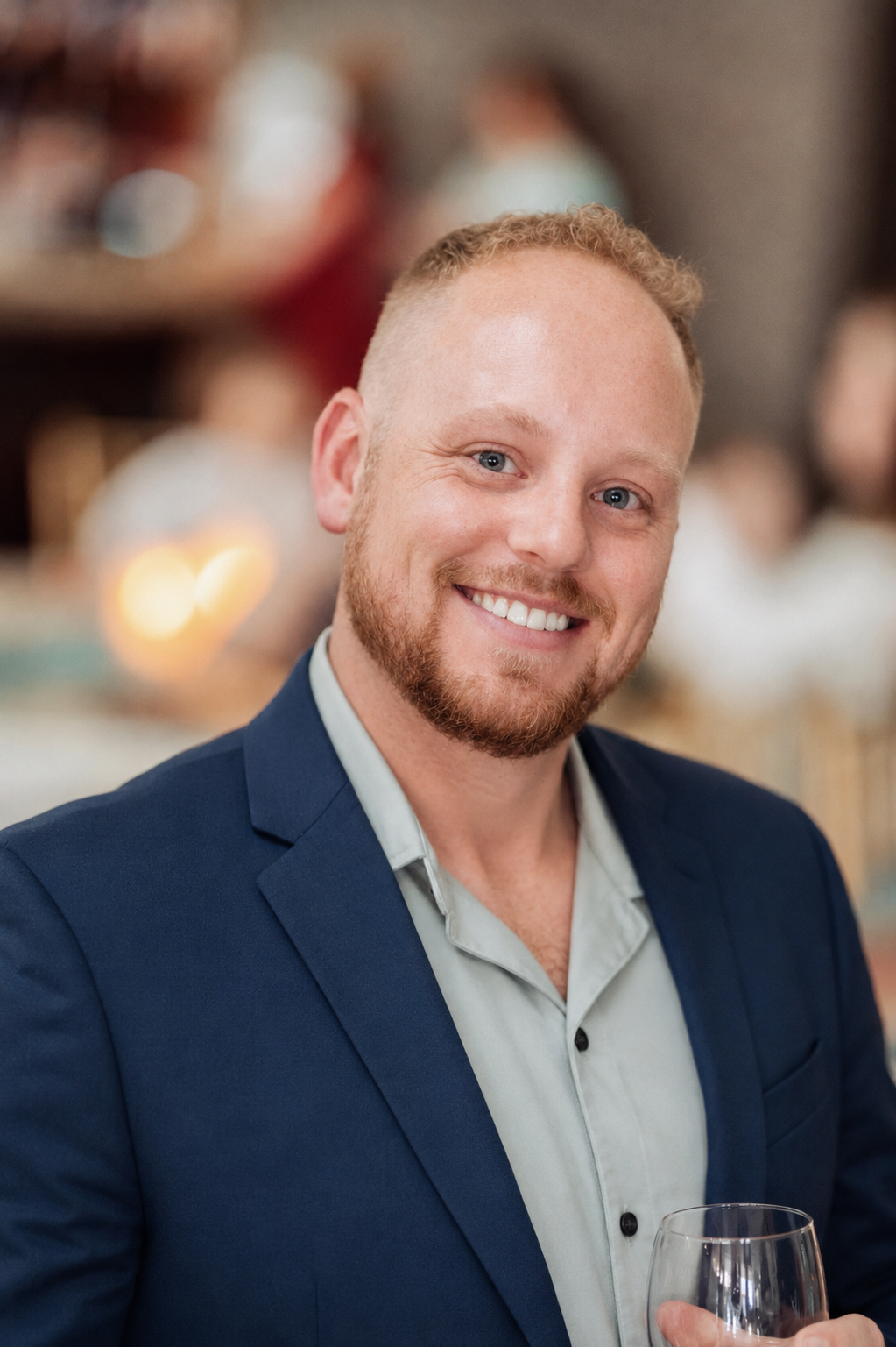 Smiling man with red beard wearing navy blazer holding wine glass at elegant indoor event.