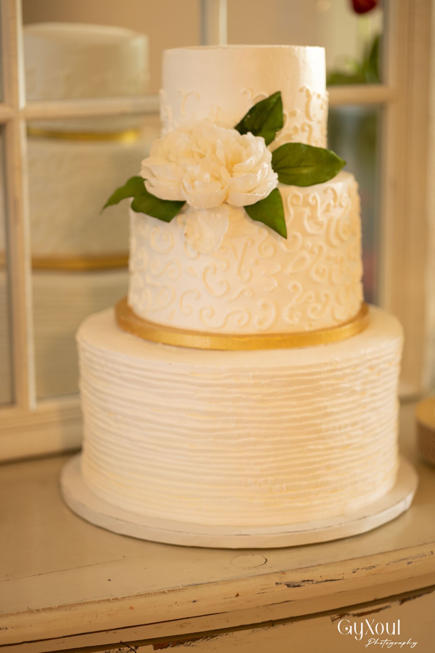 Elegant three-tier white wedding cake with gold band, scroll details, and fresh white peony flower decoration.