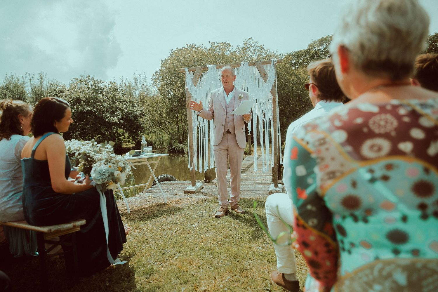 Outdoor wedding ceremony with officiant speaking to seated guests under decorative arch in garden setting.