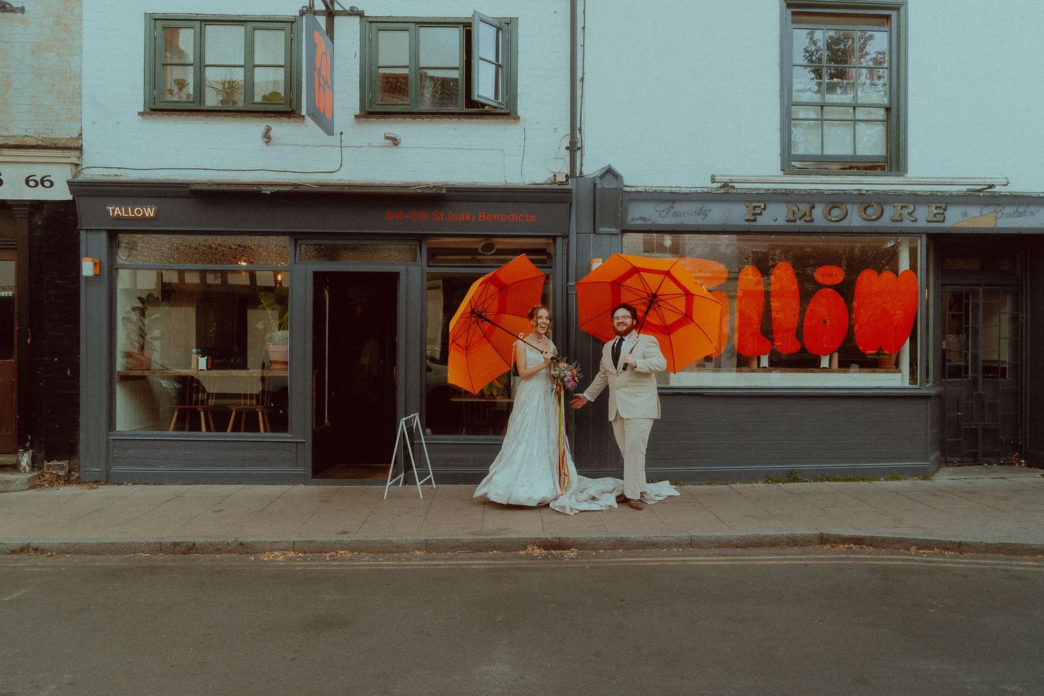 Bride and groom holding vibrant orange umbrellas outside colorful storefronts on a charming street.