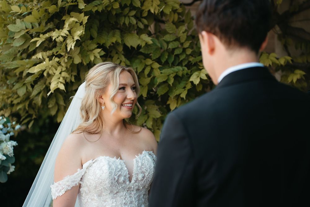 Bride in lace off-shoulder gown smiling at groom during outdoor wedding ceremony with lush green foliage backdrop.