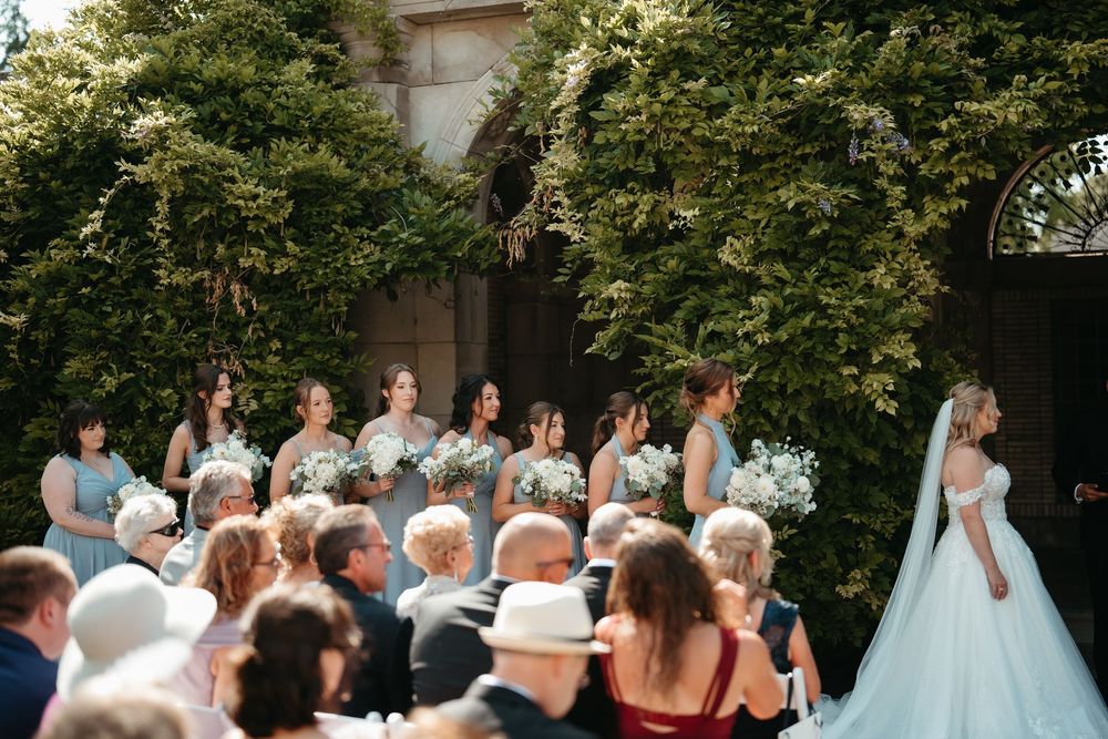 Bride in white gown stands before bridesmaids in blue dresses holding bouquets at an outdoor wedding ceremony.
