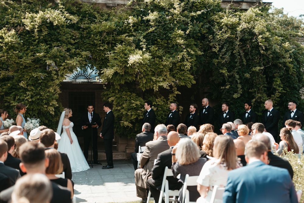 Outdoor wedding ceremony with bride and groom at the altar, groomsmen in black suits, and seated guests surrounded by lush greenery.