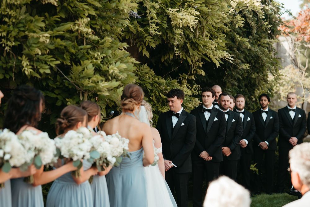 Outdoor wedding ceremony with bridesmaids in light blue gowns holding white bouquets facing groomsmen in black tuxedos.