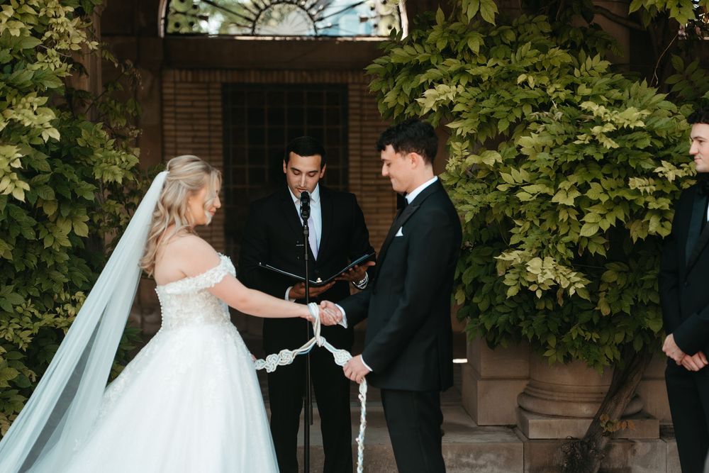 Bride and groom exchange vows outdoors with officiant, surrounded by lush greenery, in an elegant wedding ceremony.