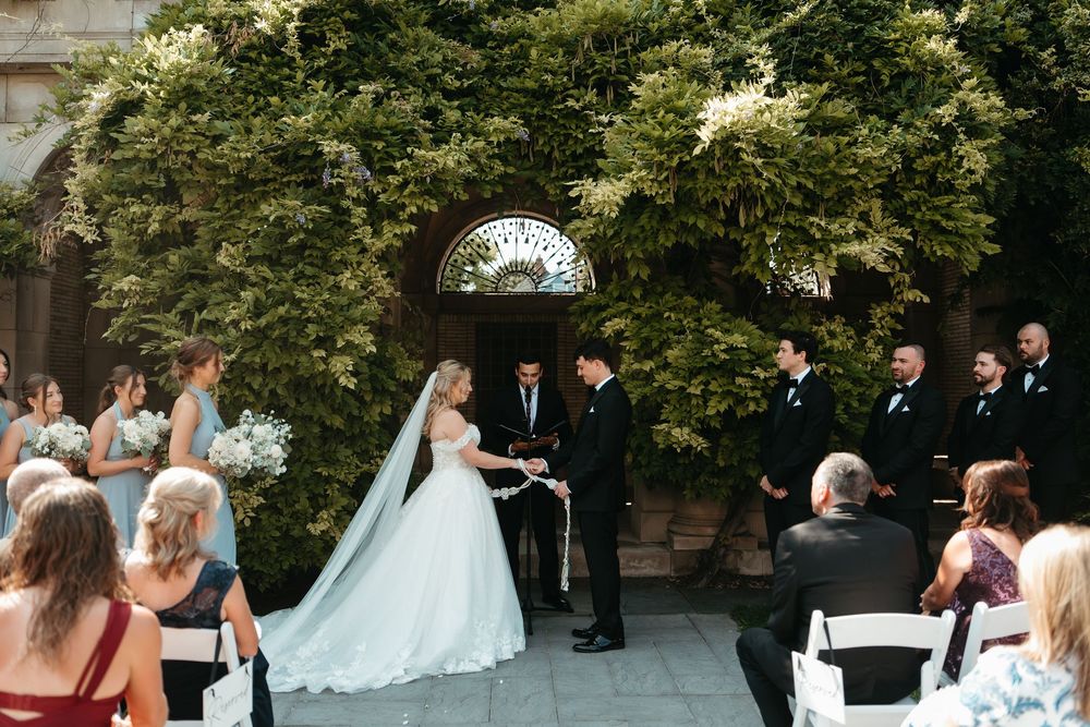 Outdoor wedding ceremony with bride in white gown and veil exchanging vows before officiant, groomsmen, and seated guests.