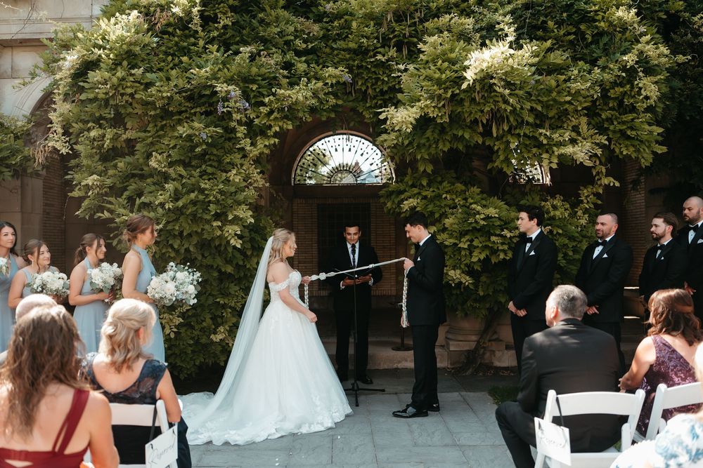 Bride and groom exchange vows at an outdoor wedding ceremony with bridesmaids and groomsmen beside a vine-covered stone building.