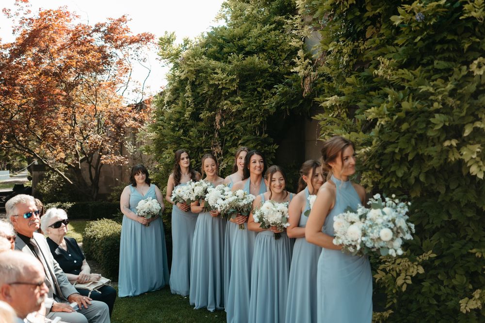 Bridesmaids in light blue gowns holding white bouquets stand outdoors at a wedding ceremony near lush green ivy walls.