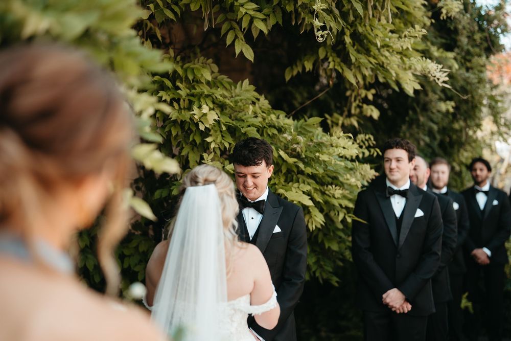 Groom in black tuxedo smiling at bride during outdoor wedding ceremony with groomsmen standing behind him.
