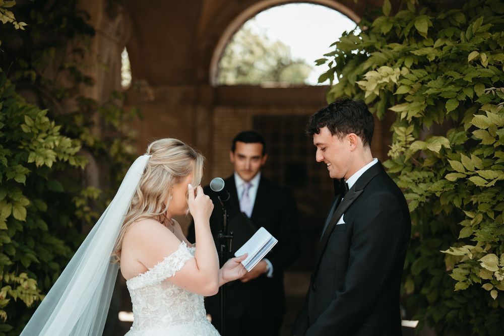 Bride reads emotional wedding vows while groom smiles during outdoor ceremony with officiant and lush greenery backdrop.