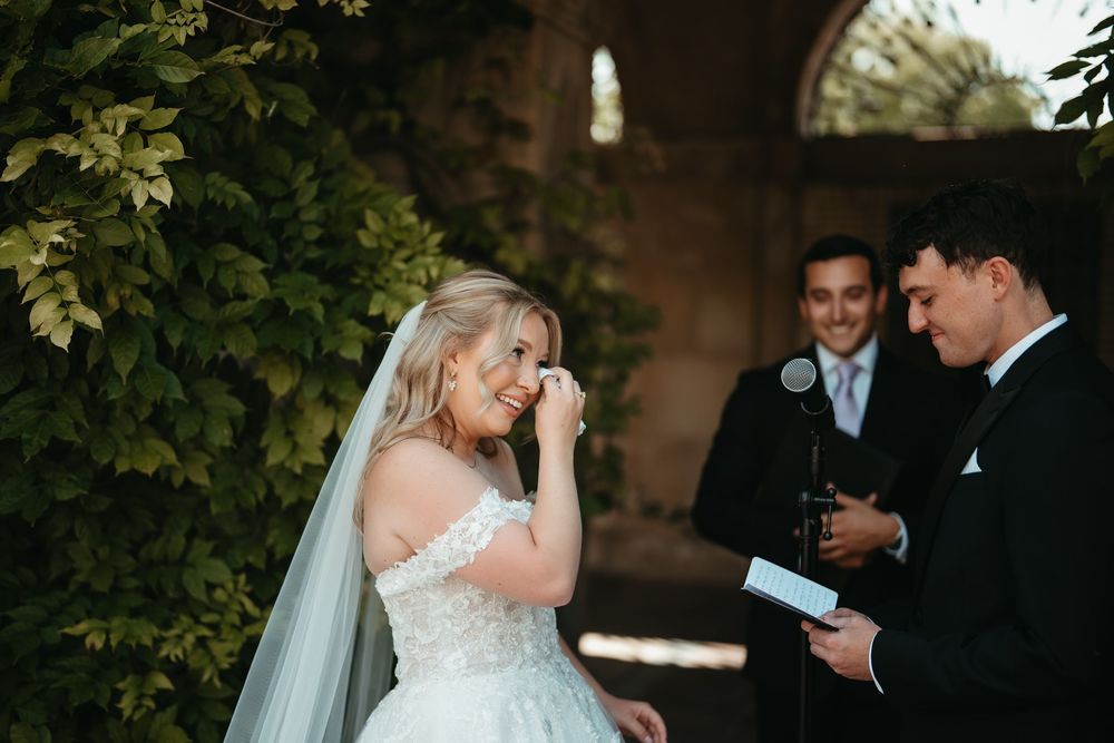 Emotional bride in off-shoulder white ballgown wipes tears during outdoor wedding ceremony while groom reads vows.