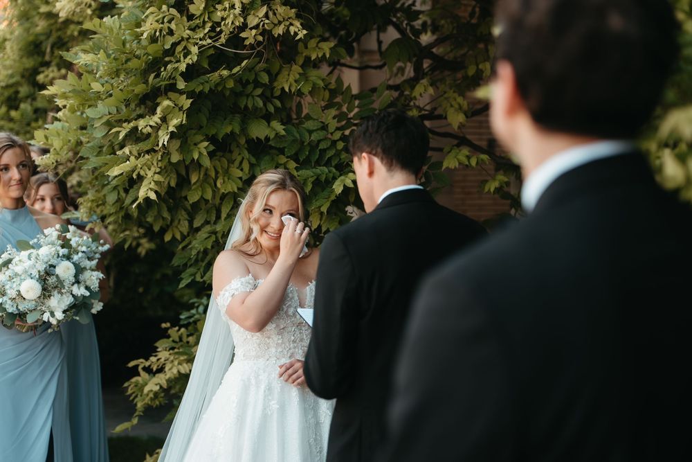 Emotional bride wiping tears during outdoor wedding ceremony with bridesmaids in blue dresses beside her.
