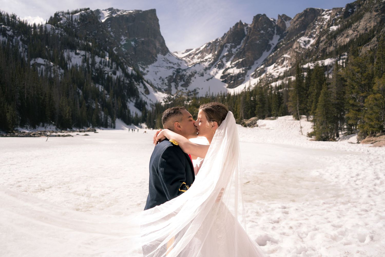 Bride and groom kissing in snowy Rocky Mountain landscape with frozen lake and pine trees behind them.