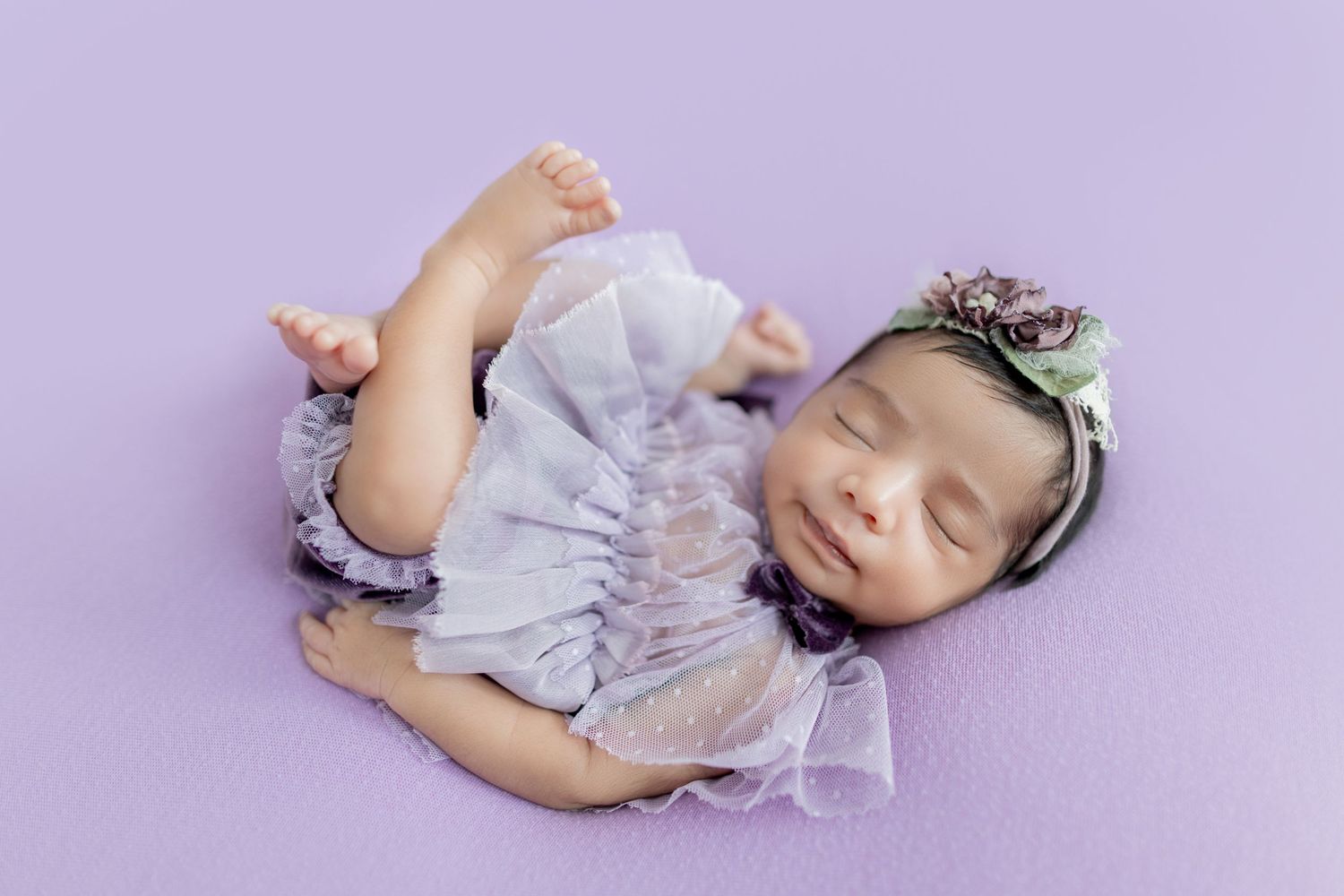 Smiling newborn baby girl in lavender ruffle dress and floral headband sleeping on purple backdrop.
