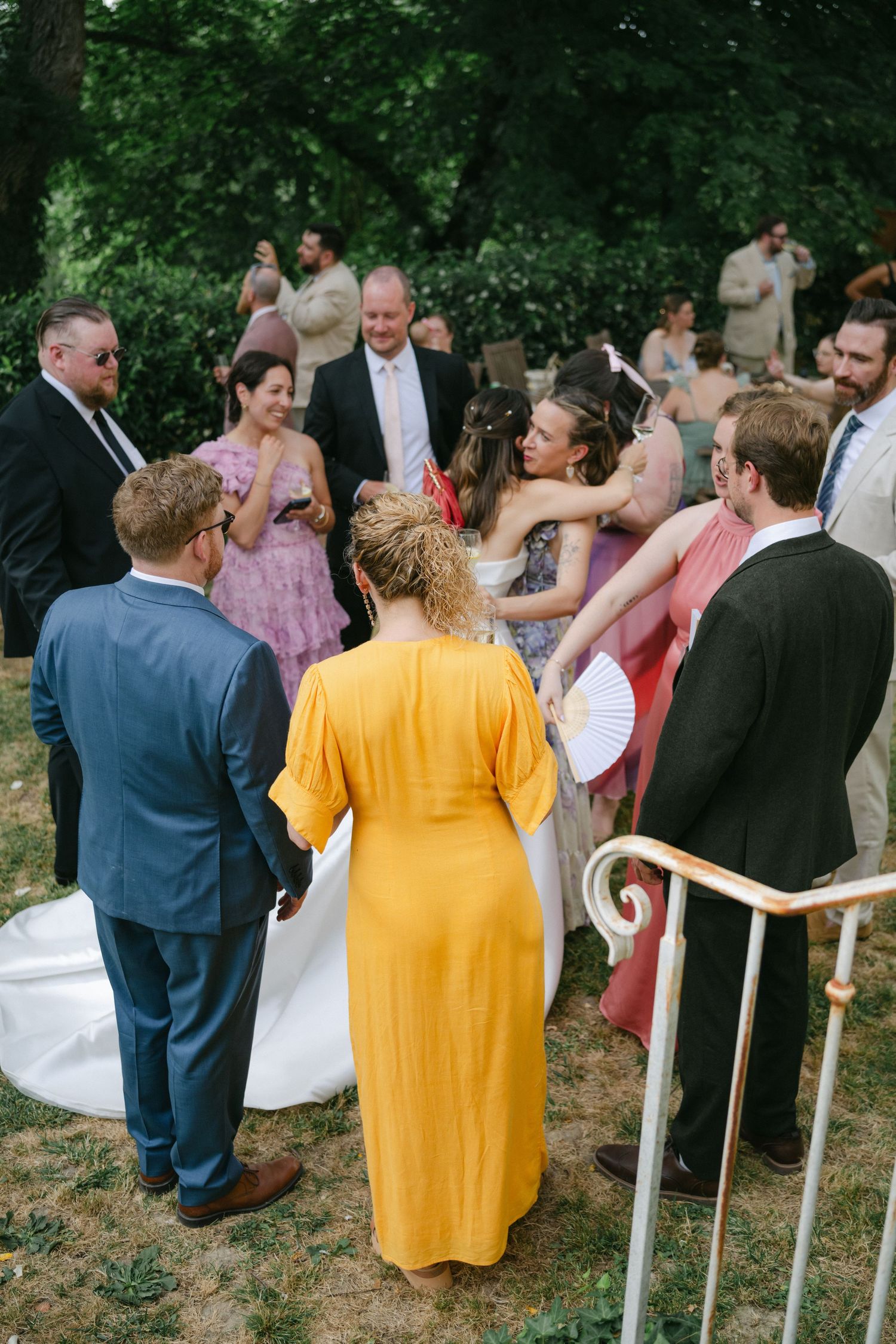 Wedding guests gathered outdoors, bride in white gown with woman in yellow dress celebrating near garden steps.