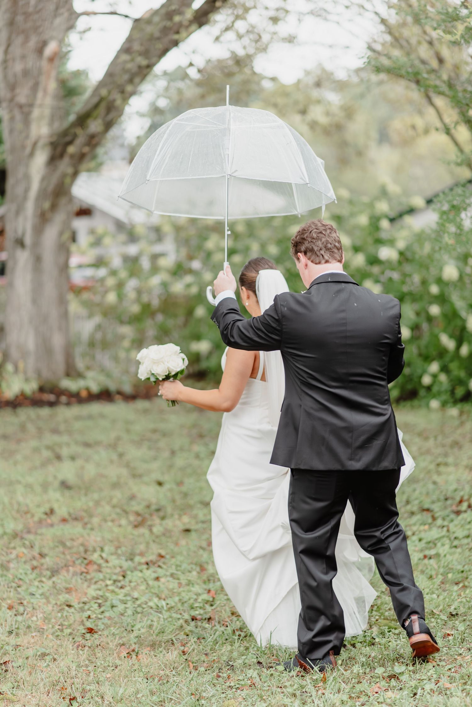 Bride and groom pose with fluffy dog under willow tree on green lawn, bride holding white bouquet in elegant wedding attire.