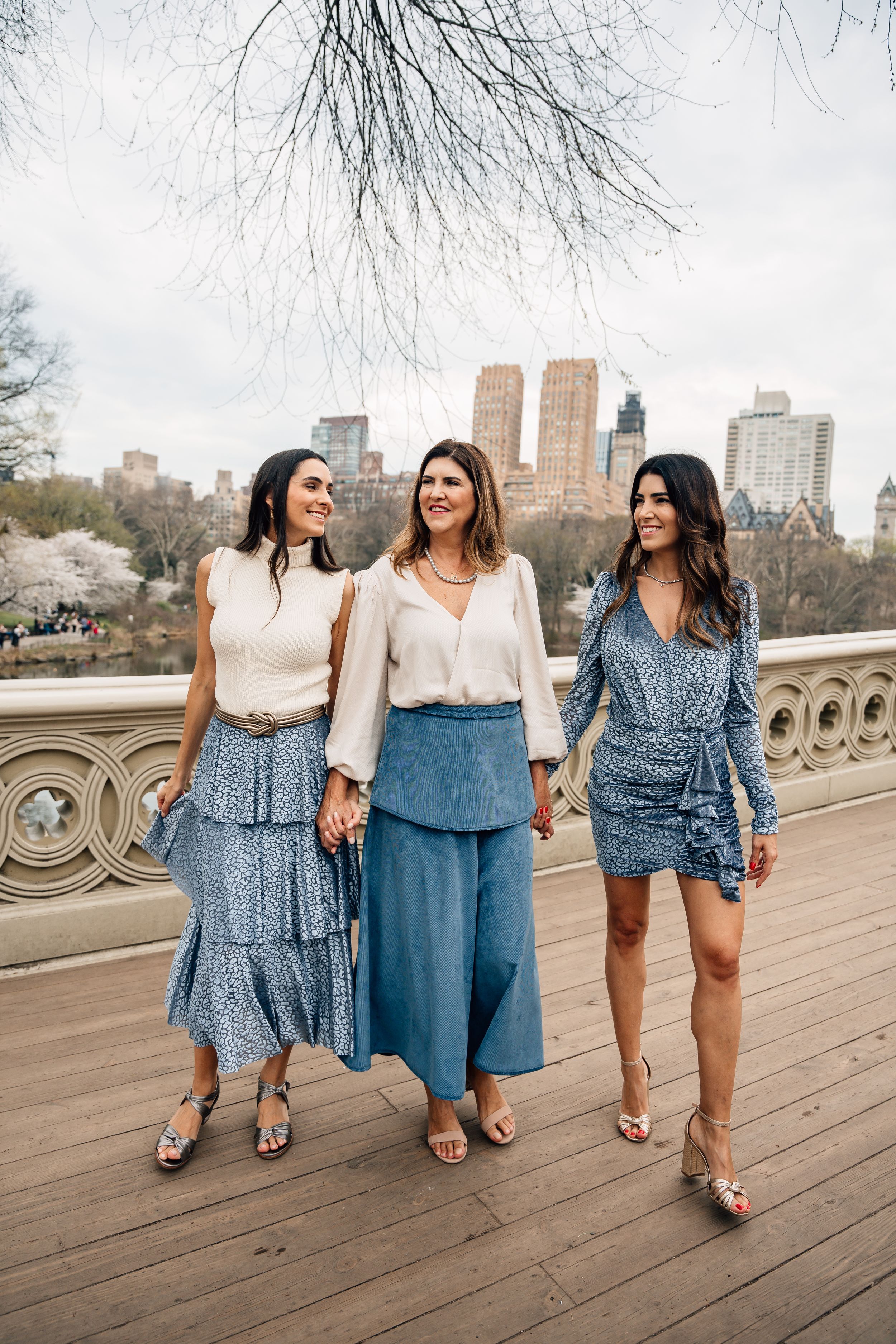 Three women in blue outfits posing on a bridge in Central Park with NYC skyline in the background.