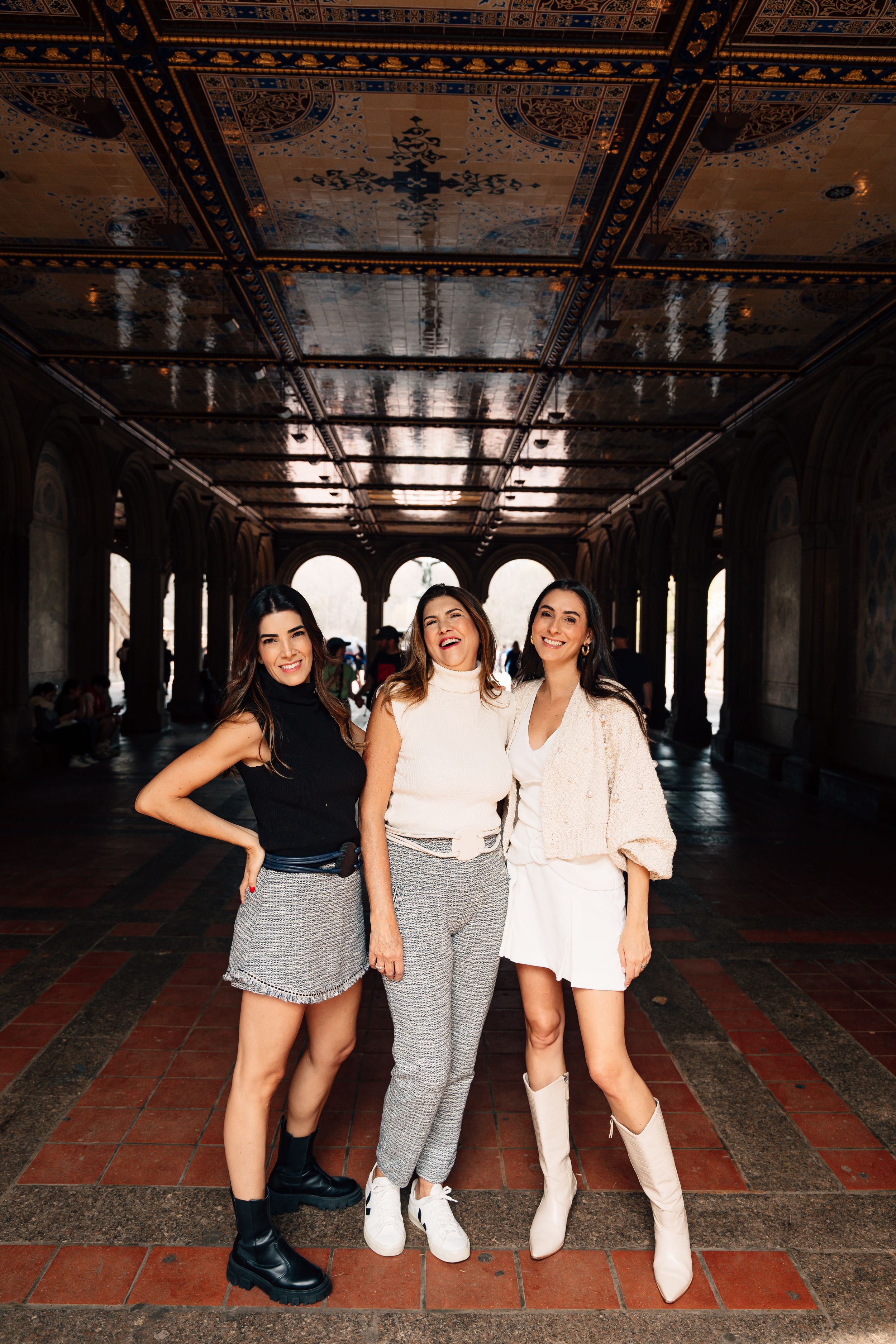 Three stylish women posing together in Bethesda Terrace arcade, Central Park, NYC, wearing trendy outfits.