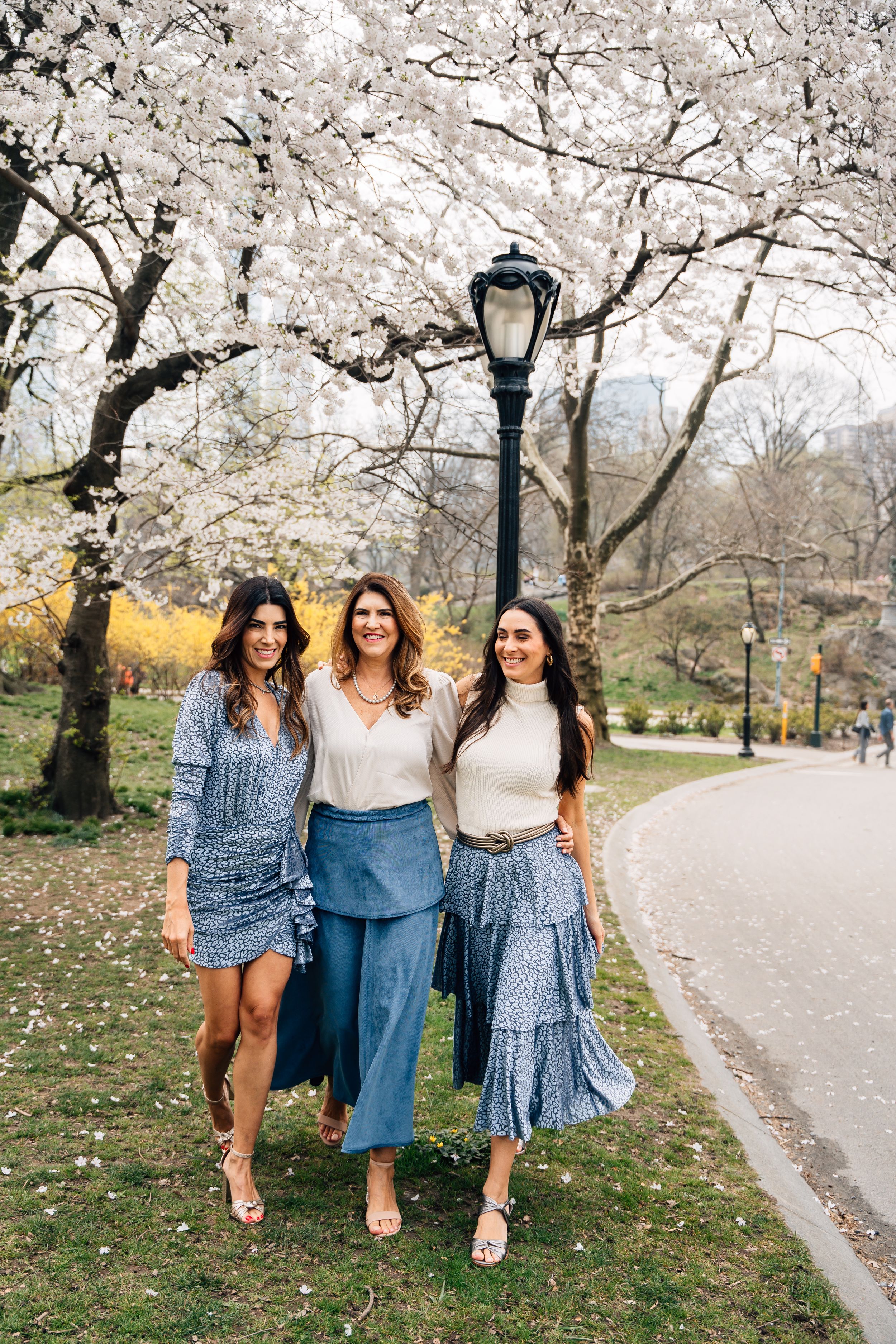 Three women in blue outfits smile under blooming cherry blossom trees in Central Park on a spring day.
