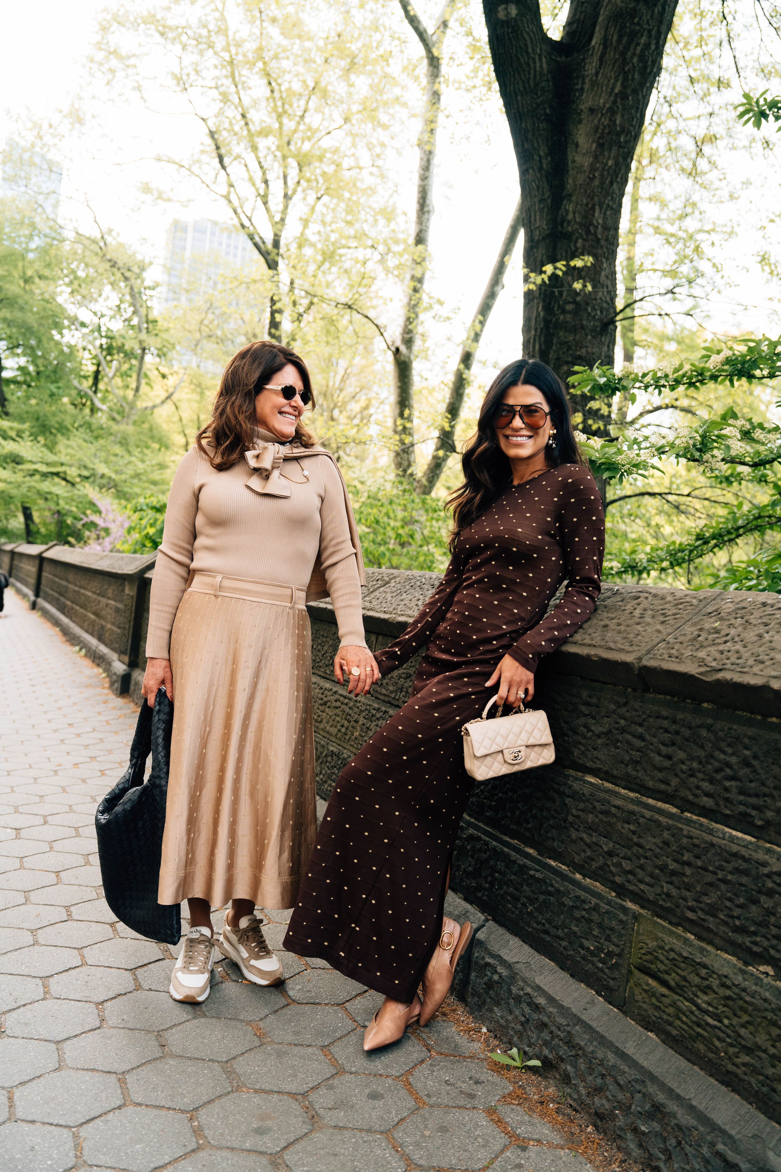 Two stylish women in elegant outfits pose in Central Park, wearing sunglasses and carrying designer handbags.