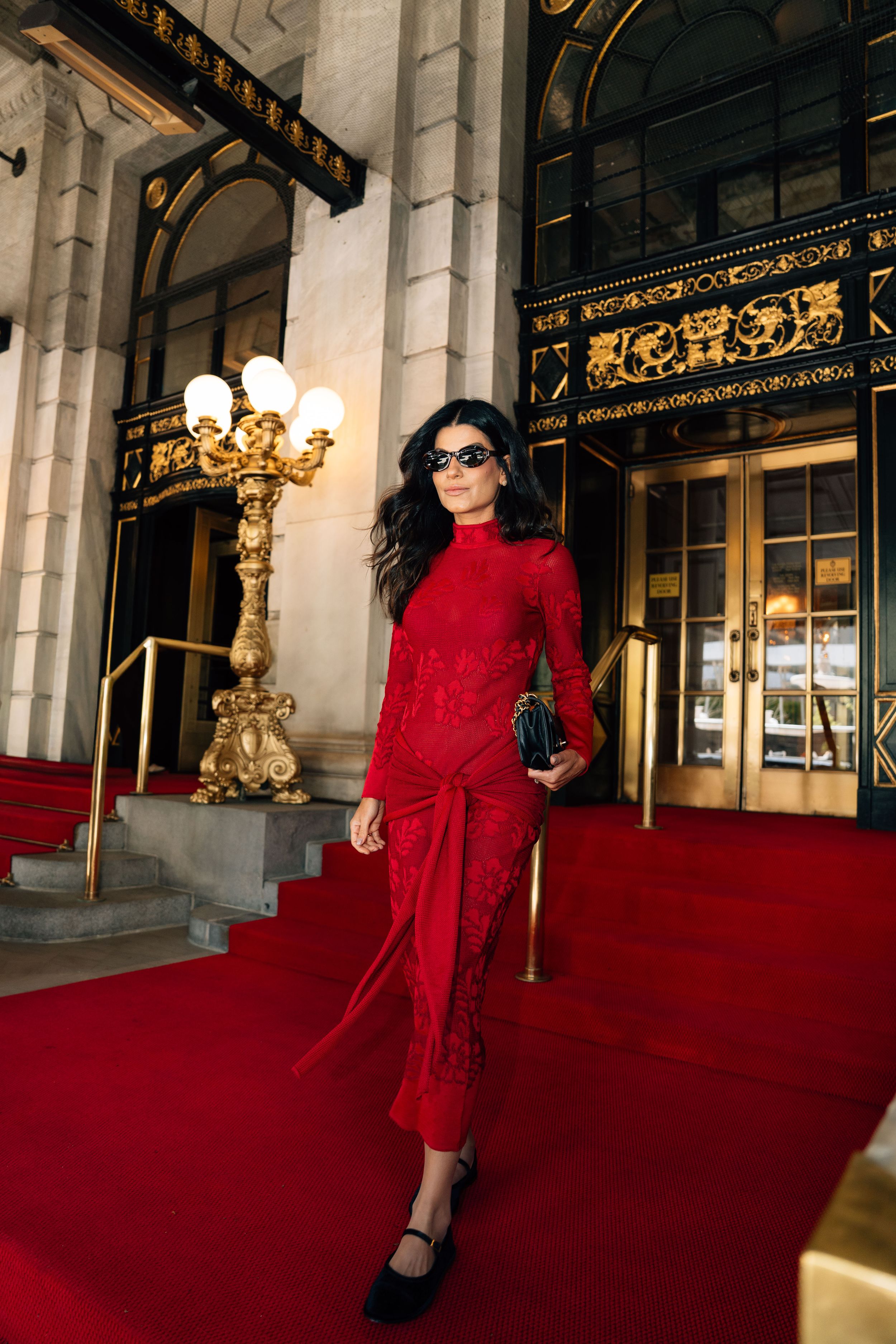 Woman in red lace midi dress and sunglasses poses on red carpet steps outside elegant gold-trimmed building entrance.