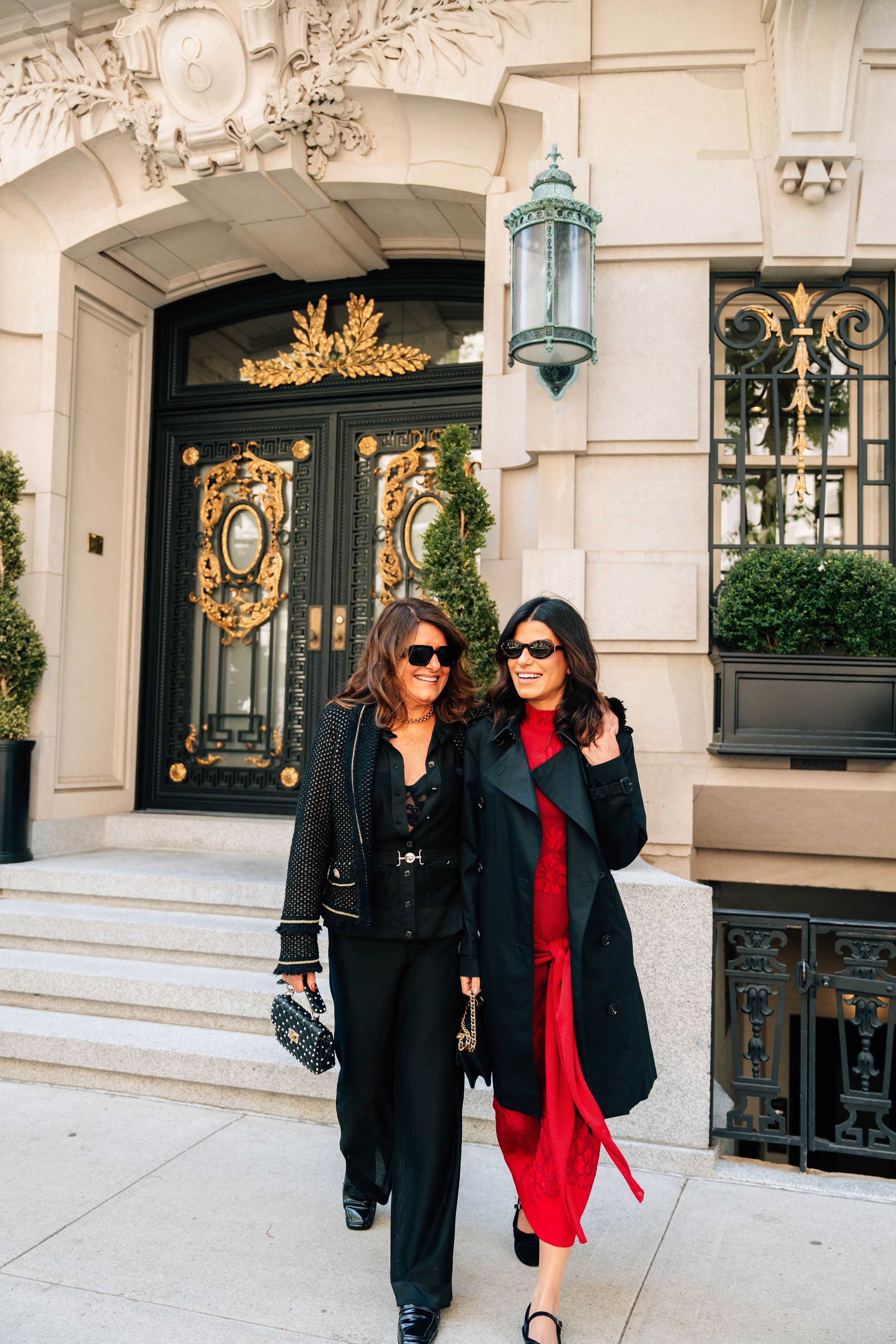 Two stylish women in black and red outfits walk outside an elegant building with ornate gold doors.