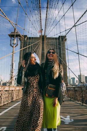 Two stylish friends enjoying a sunny day on the Brooklyn Bridge walkway.