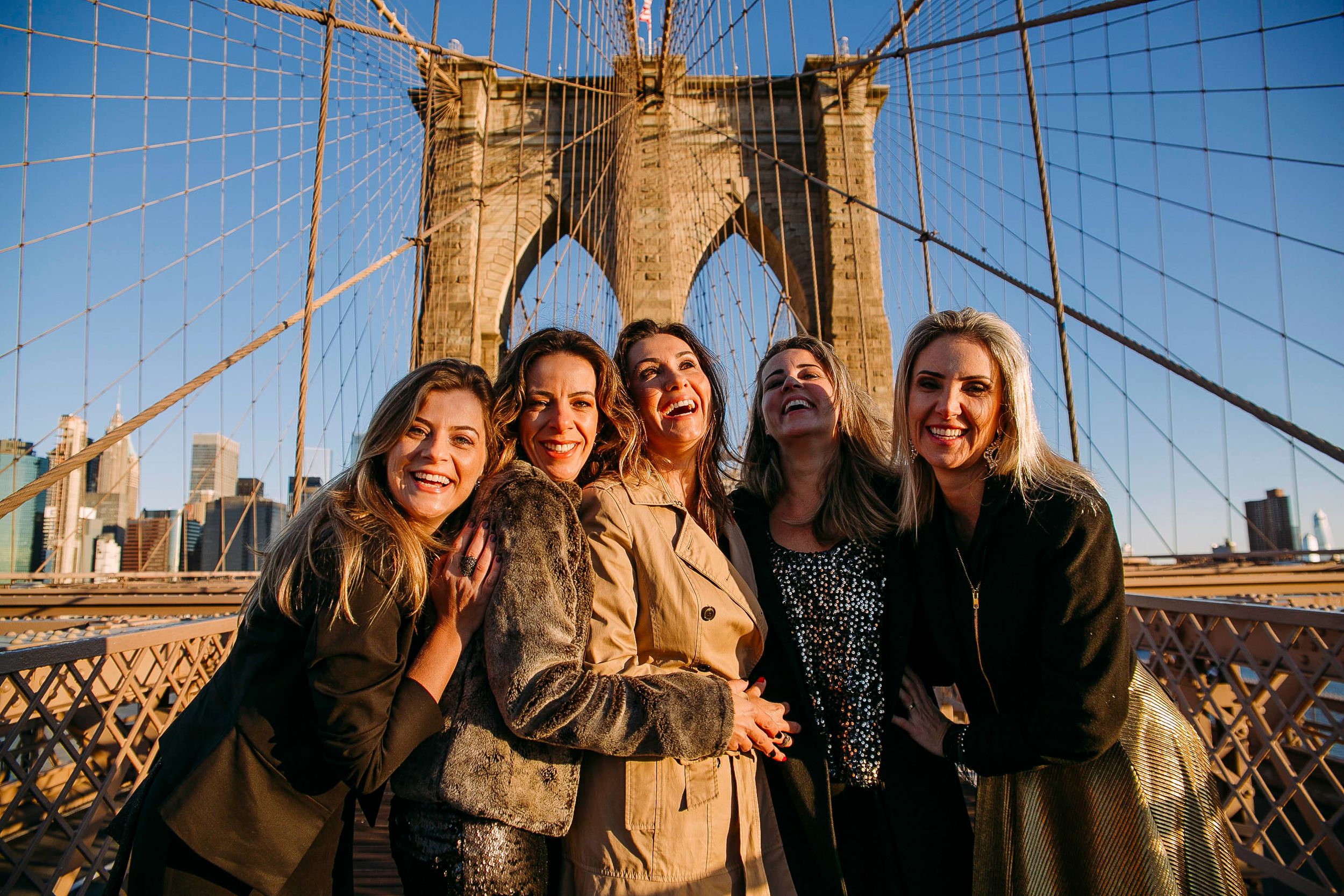 Friends sharing joyful moment on Brooklyn Bridge with NYC skyline visible behind.