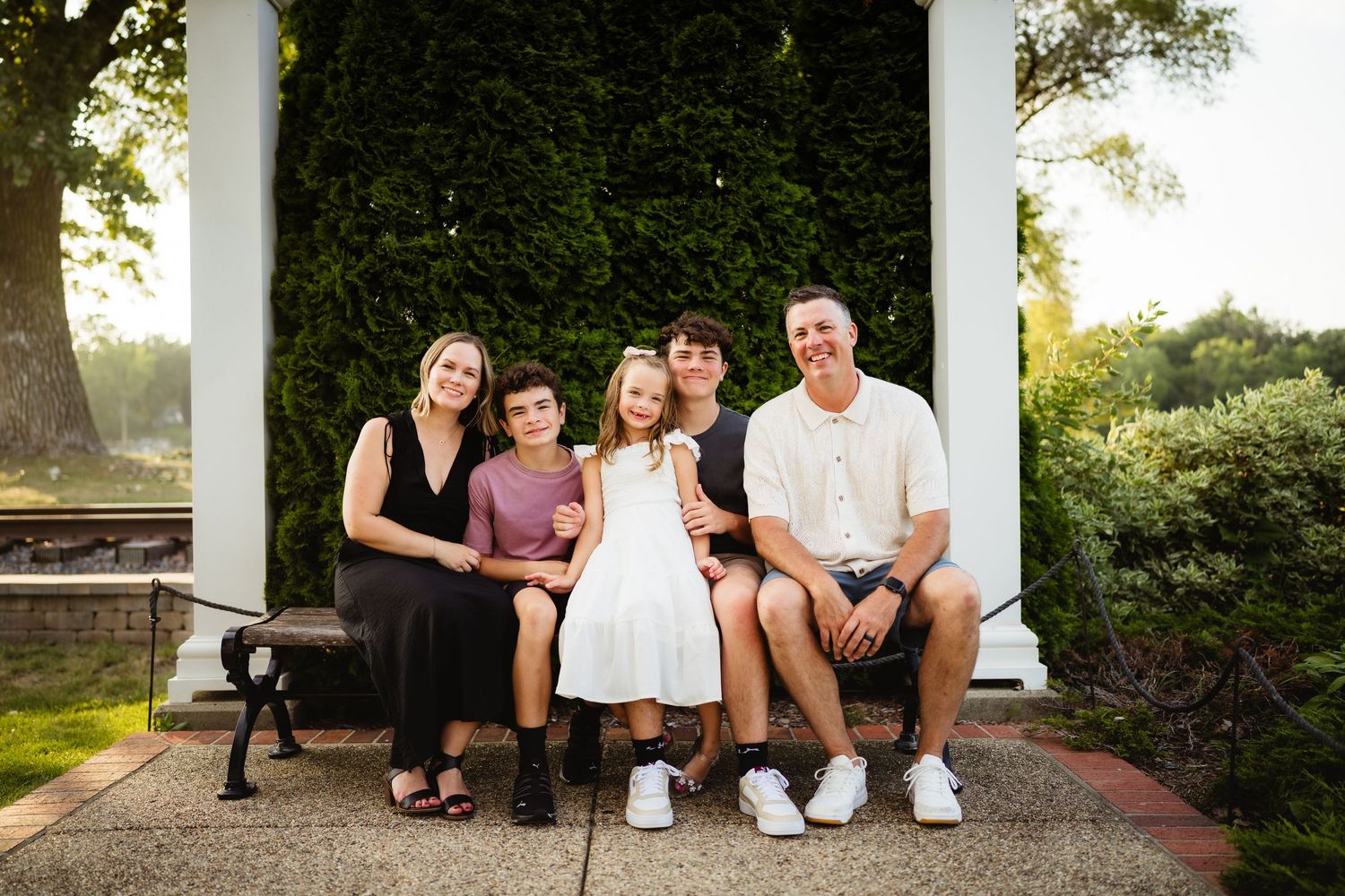 Family of five posing on a bench under a white pergola surrounded by lush greenery during a golden hour outdoor photo session.