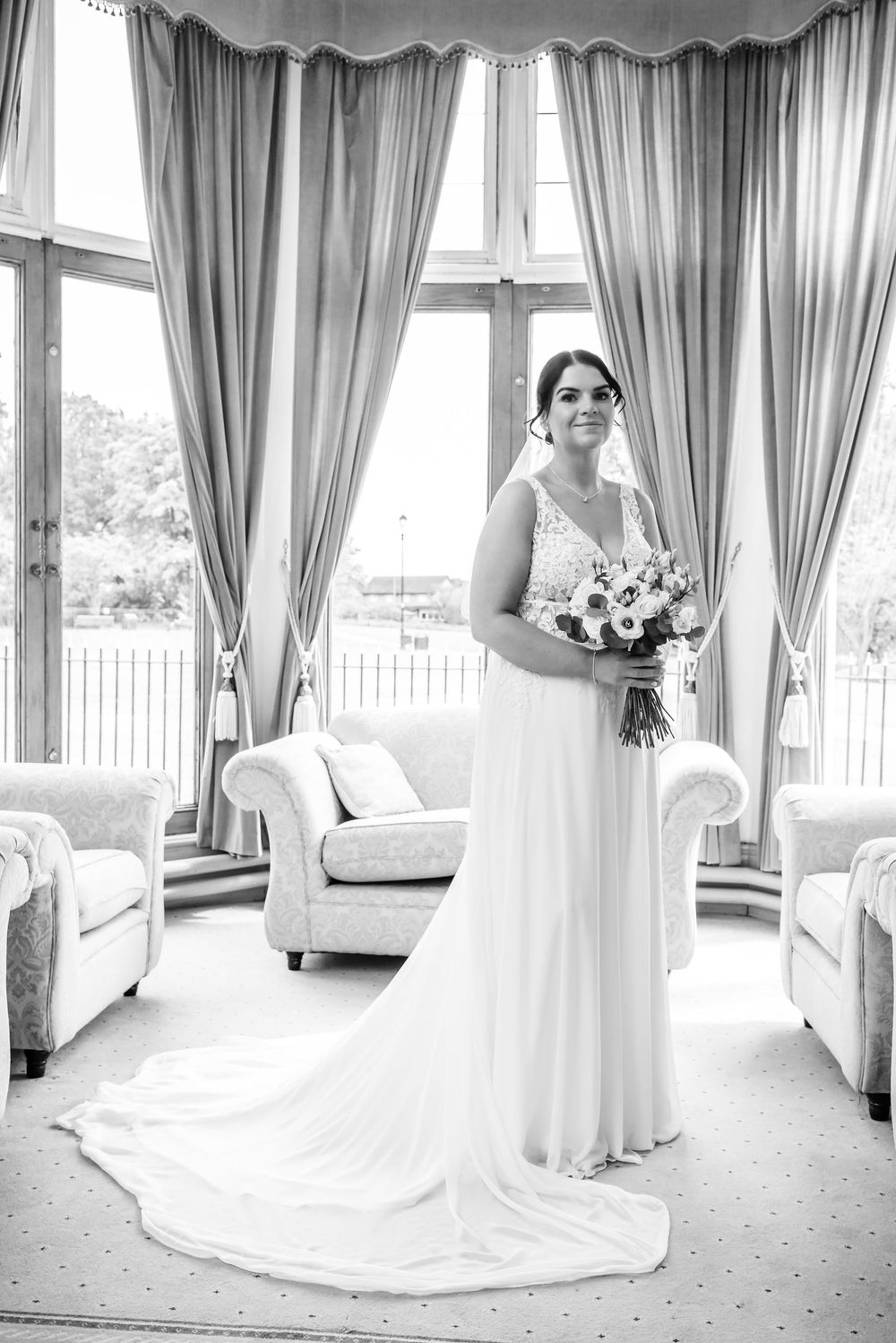 Black and white portrait of a bride in a flowing white gown holding a bouquet by elegant draped windows.