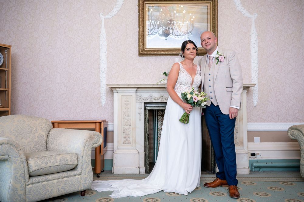 Bride and groom posing together in elegant room with fireplace, bride holding white bouquet, groom in beige suit.