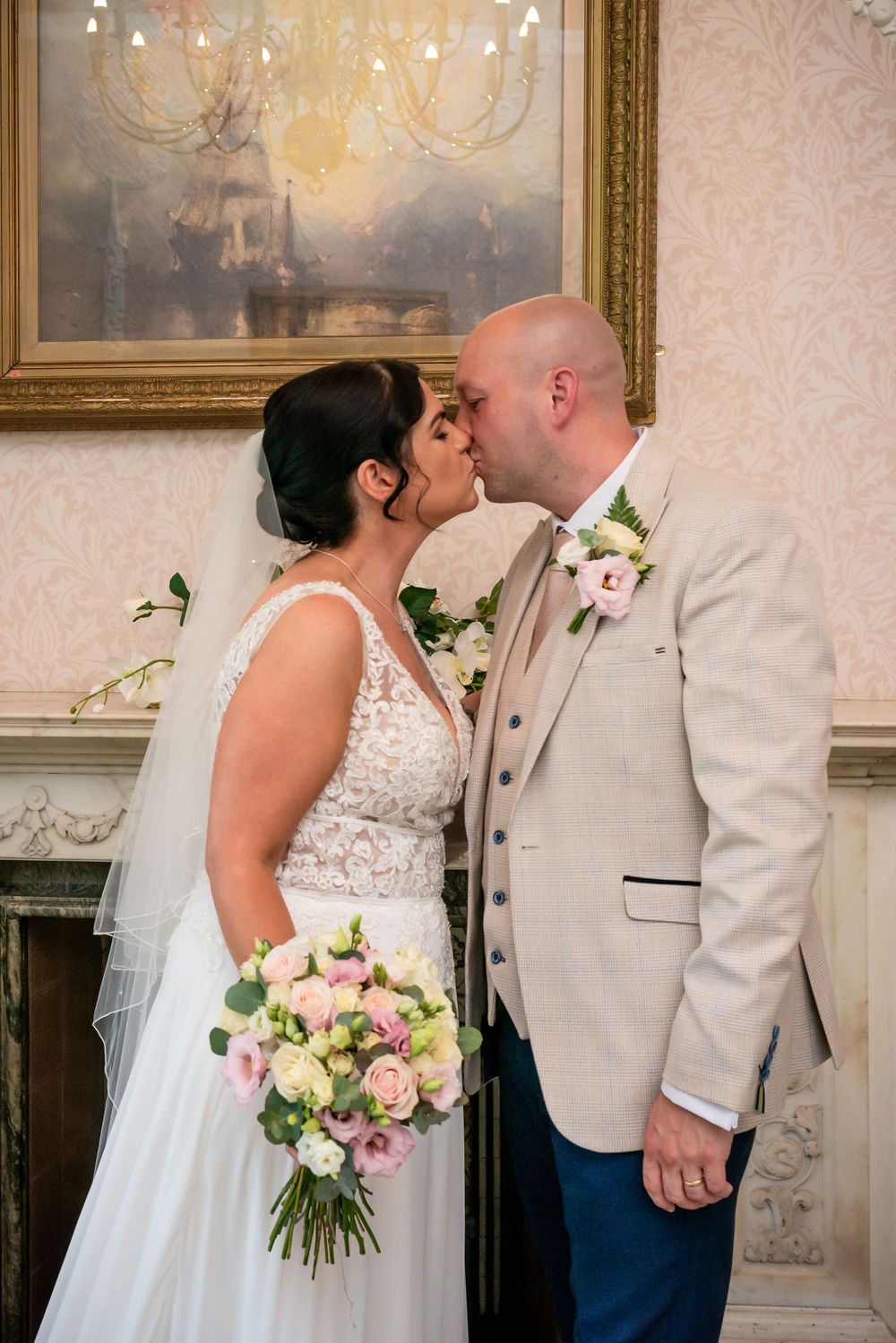 Bride and groom sharing a romantic kiss indoors, bride holding pink bouquet in lace wedding dress with veil.