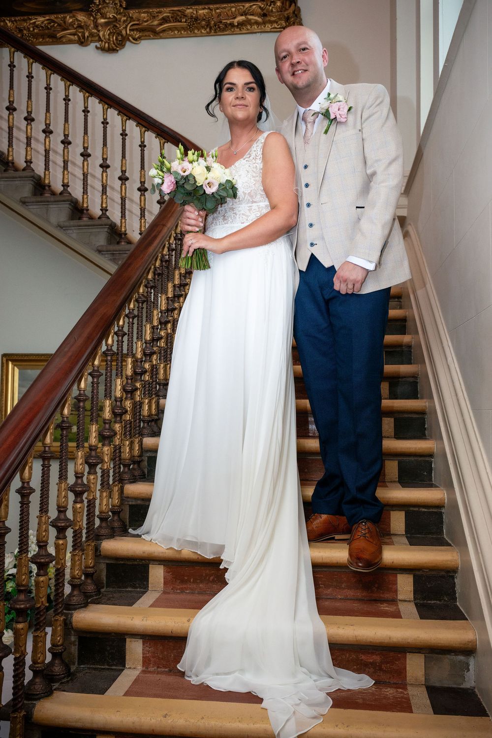 Bride and groom posing elegantly on a grand staircase, bride holding a bouquet in a white gown.