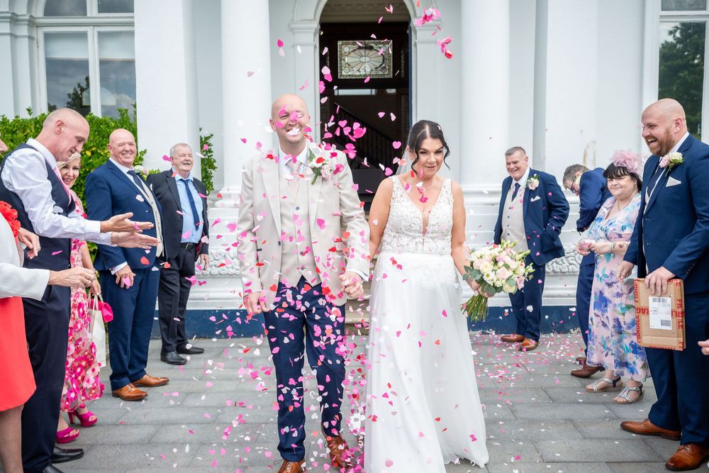 Bride and groom walk through confetti shower surrounded by cheering guests outside elegant white venue.