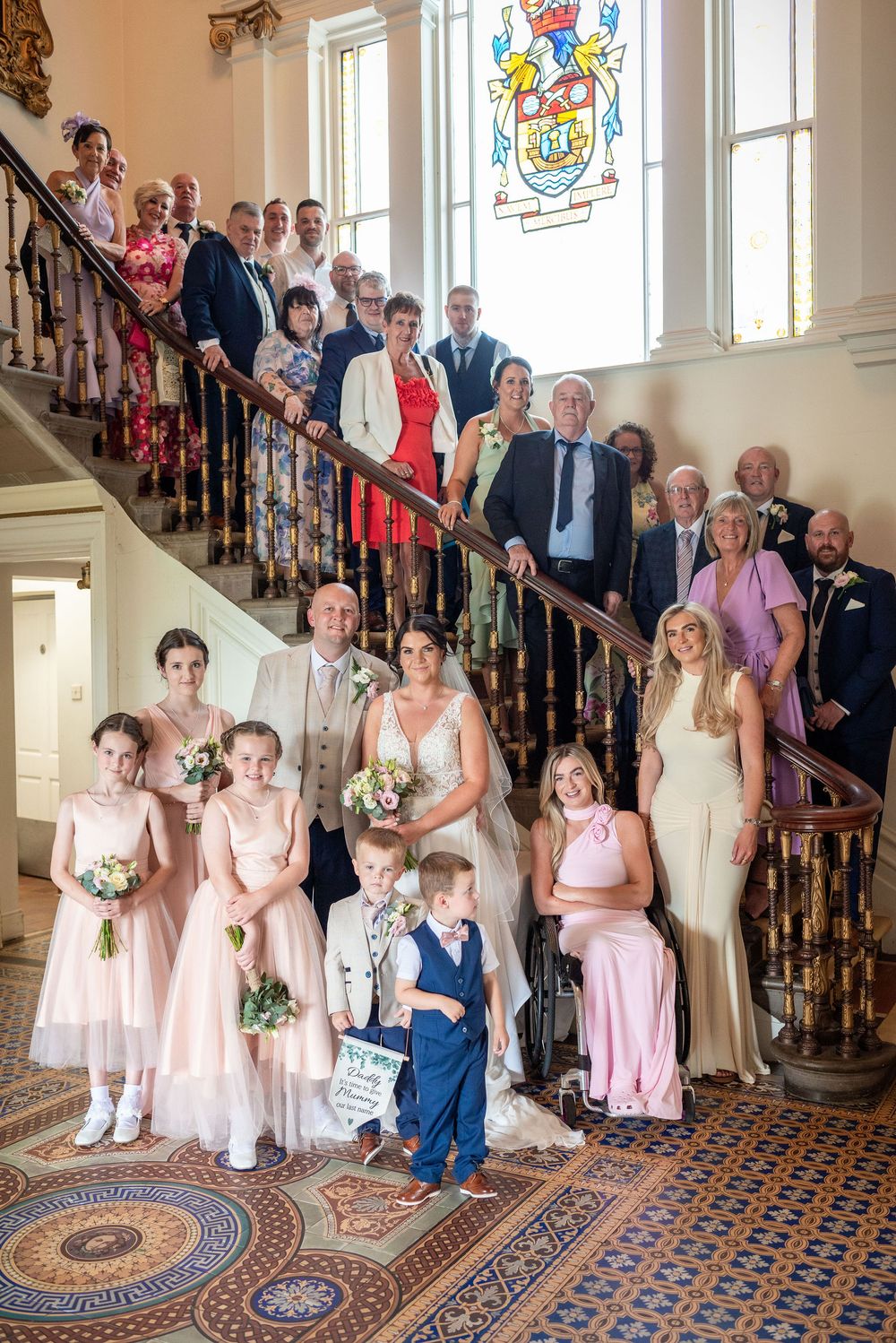 Wedding party group photo on ornate staircase with stained glass window, including bridesmaids and guests.