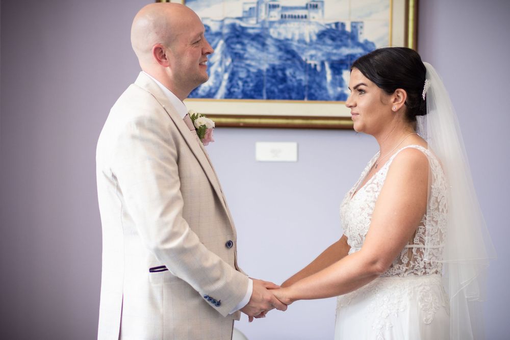 Bride and groom holding hands during wedding ceremony, wearing white lace gown and beige suit.