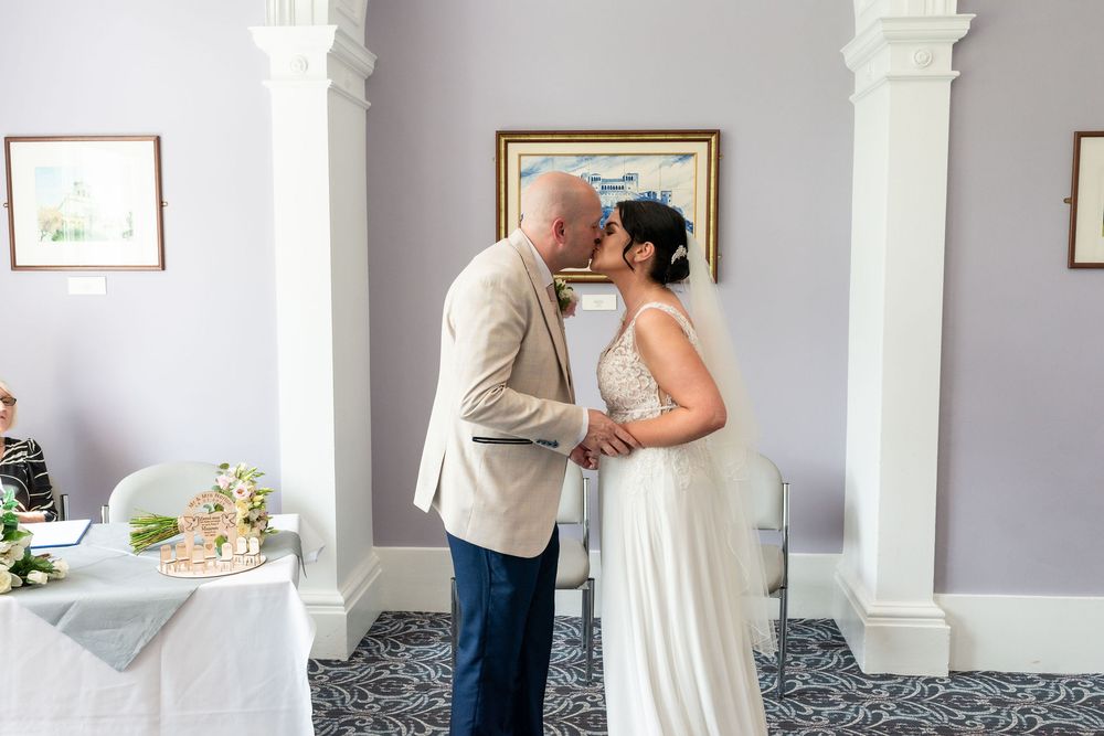 Bride and groom share a first kiss during intimate wedding ceremony in elegant white room with columns.