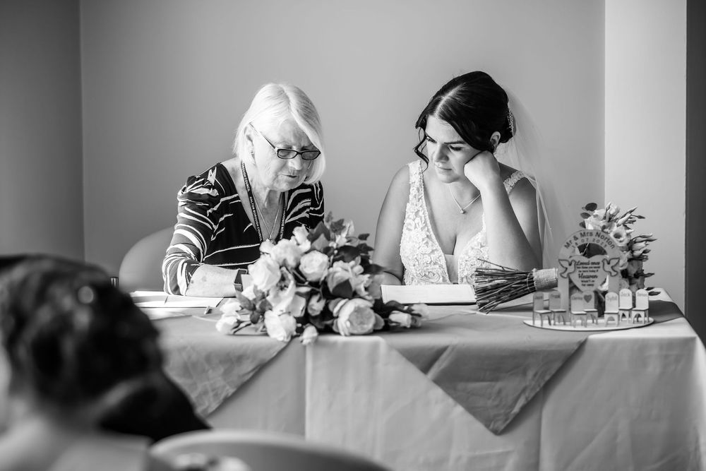 Black and white photo of a bride and older woman reviewing documents at a decorated wedding table with flowers.