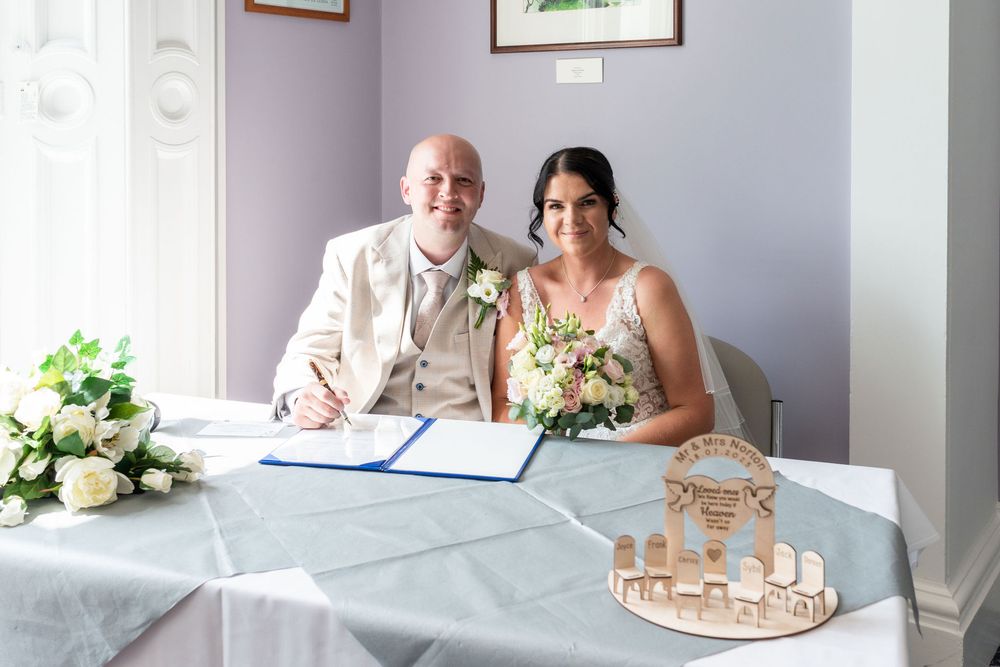 Newlywed couple signing wedding register at table with flowers and decorative centerpiece in elegant room.