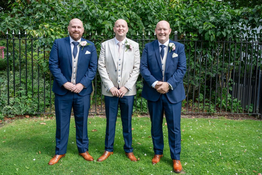 Three groomsmen in navy suits pose outdoors, with the groom in a light grey suit and white boutonniere in the center.