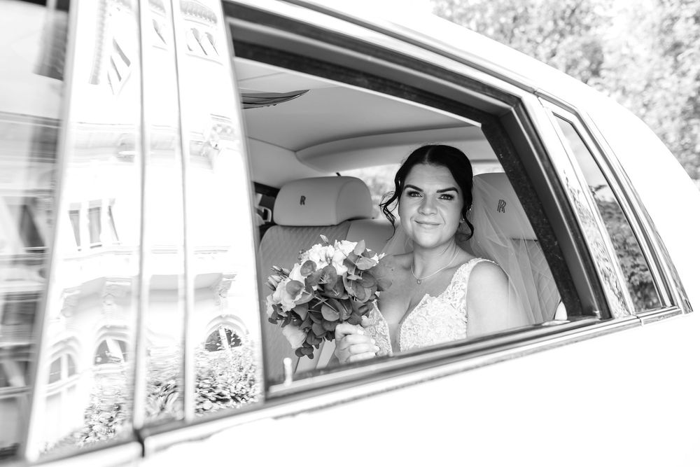 Black and white photo of a smiling bride holding a bouquet, seated in a luxury car on her wedding day.