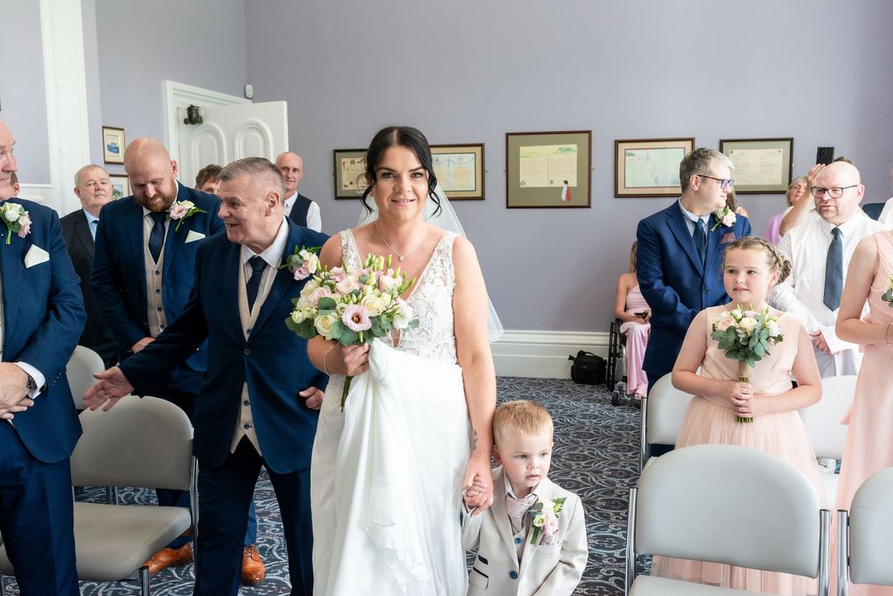 Bride walks down the aisle with young children holding bouquets, guests seated in blue chairs at elegant wedding venue.