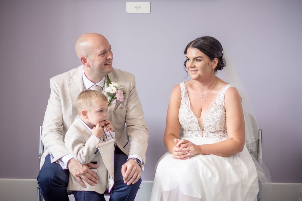 Bride and groom in cream suit sit with young child during wedding ceremony in a softly lit venue.