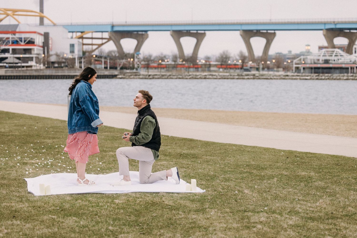 Man proposes on one knee to woman by riverside on grassy lawn with bridge in background.